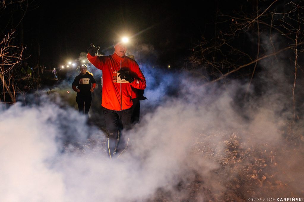 Uczestnik wydarzenia biegnie nocną porą leśną ścieżką. Jest ubrany w czerwoną kurtkę, na głowie ma latarkę - czołówkę. Za nim widocznych jest więcej biegnących osób.