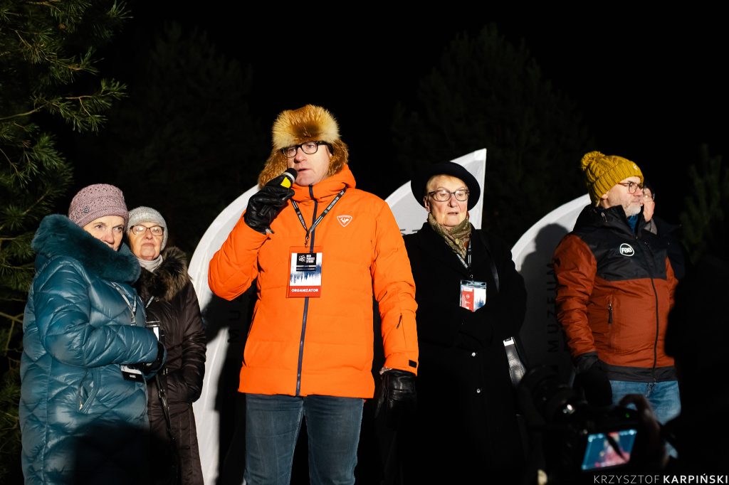 Prof. Wojciech Śleszyński, dyrektor Muzeum Pamięci Sybiru przemawia do mikrofonu. Ubrany jest w pomarańczową puchową kurtkę oraz futrzaną czapkę. Obok niego stoją Sybiraczki oraz organizatorzy Biegu Pamięci Sybiru.