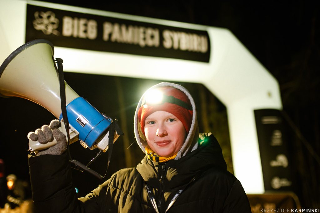 Młody chłopak trzyma uniesiony do góry megafon, na głowie ma świecącą latarkę czołową. W tle napis Bieg Pamięci Sybiru.