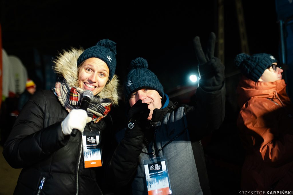 Dwie uśmiechnięte osoby pozują do zdjęcia. Są ubrane w zimowe kurtki i czapki. Jedna z osób trzyma w ręku mikrofon, druga osoba wyciąga w stronę fotografa rękę której palce układają się w literę V.