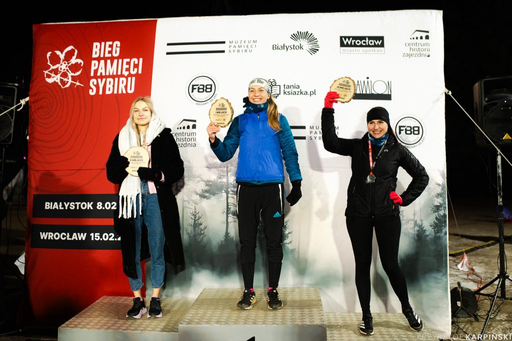 Trzy uczestniczki wydarzenia "Bieg Pamięci Sybiru" stoją przed banerem z logo i nazwami miast oraz sponsorów. Każda z nich trzyma drewnianą tabliczkę z napisem "I MIEJSCE", "II MIEJSCE" lub "III MIEJSCE", co sugeruje, że są to zwyciężczynie biegu. Są ubrane w zimowe stroje, w tym kurtki i czapki.