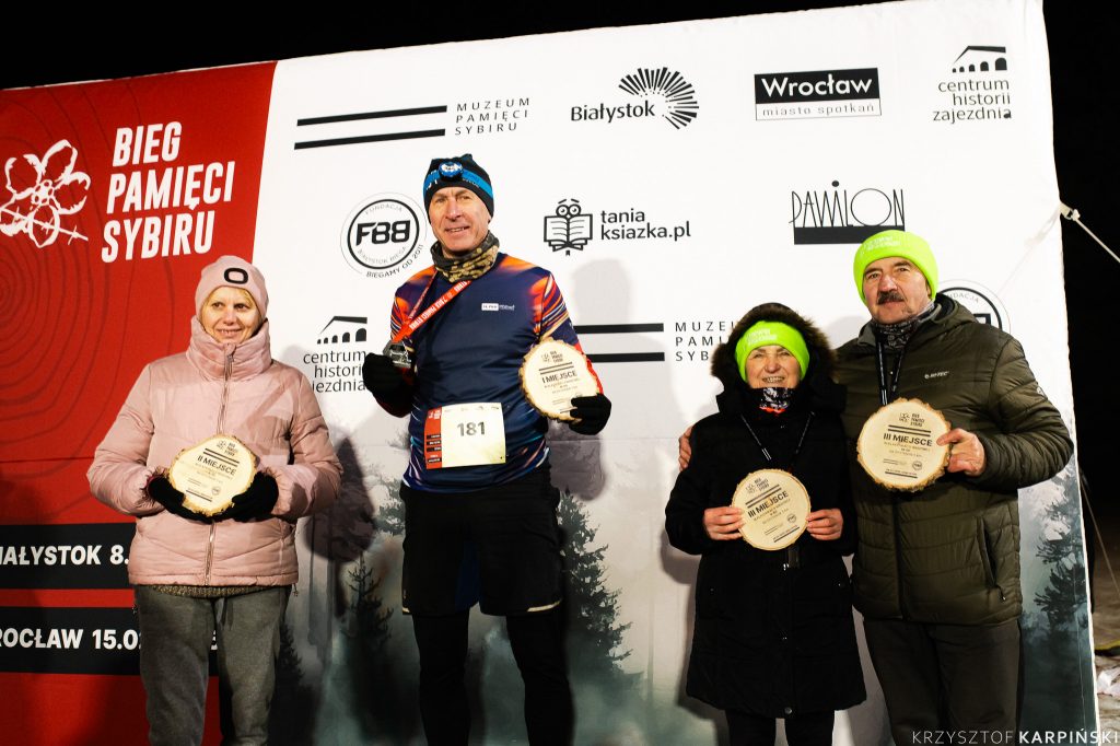 Czworo uczestników wydarzenia "Bieg Pamięci Sybiru" stojących przed banerem z logo i nazwami miast oraz sponsorów. Każdy z nich trzyma drewnianą tabliczkę z napisem "I MIEJSCE", "II MIEJSCE" lub "III MIEJSCE", co sugeruje, że są to zwycięzcy biegu. Są ubrani w zimowe stroje, w tym kurtki i czapki.