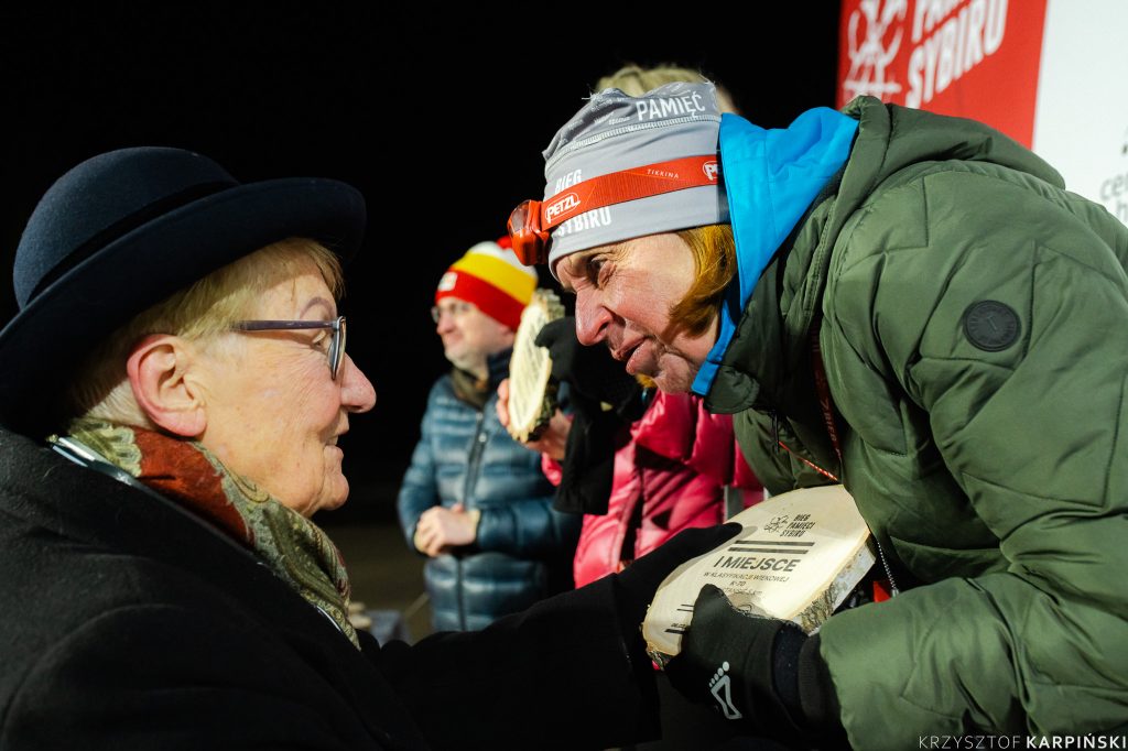 Starsza osoba w kapeluszu i w zimowym płaszczu wręcza medal kobiecie na podium.