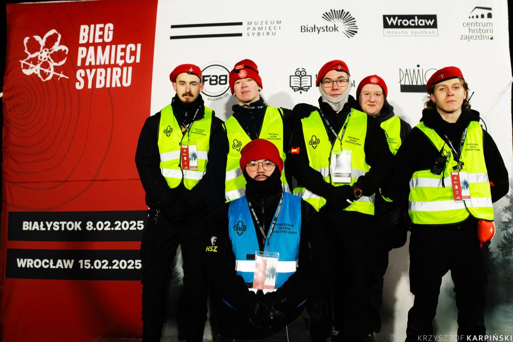 Grupa harcerzy w czarnych strojach i żółtych kamizelkach odblaskowych oraz czerwonych beretach stoi na tle baneru informującego o Biegu Pamięci Sybiru.