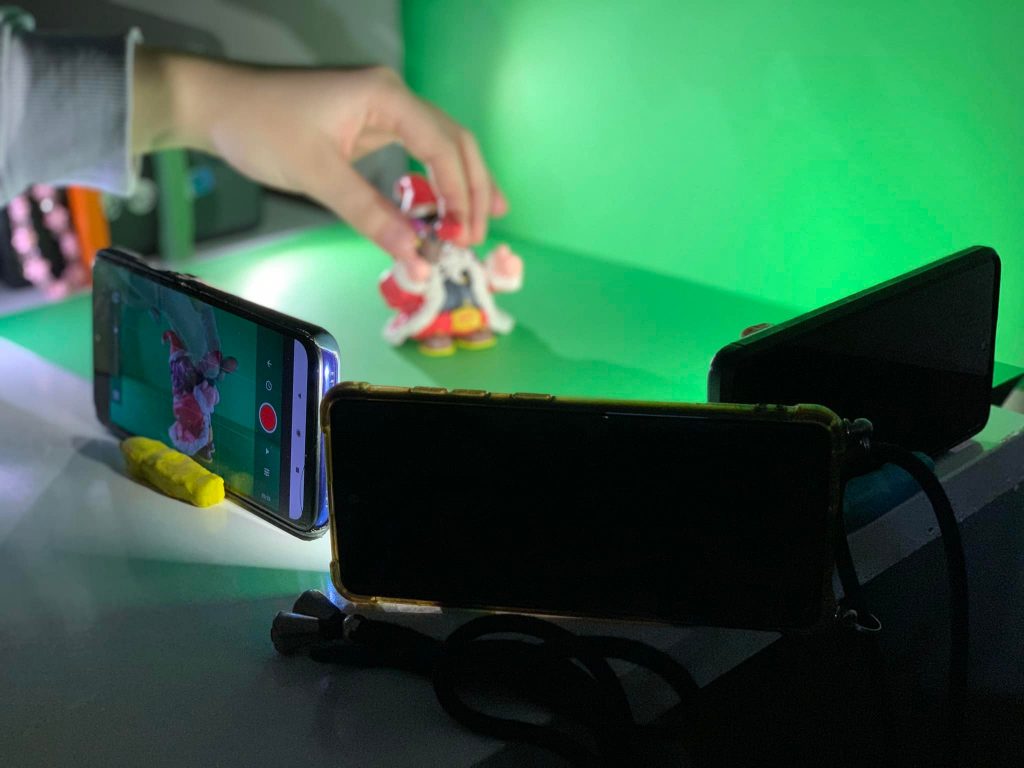 Na zdjęciu widać ustawienie do nagrywania animacji poklatkowej. Na stole znajduje się figurka w kolorowym stroju, trzymana przez uczestnika warsztatów. Dwa telefony komórkowe są ustawione na statywach, jeden z nich nagrywa scenę, co widać na ekranie, gdzie wyświetlana jest figurka.