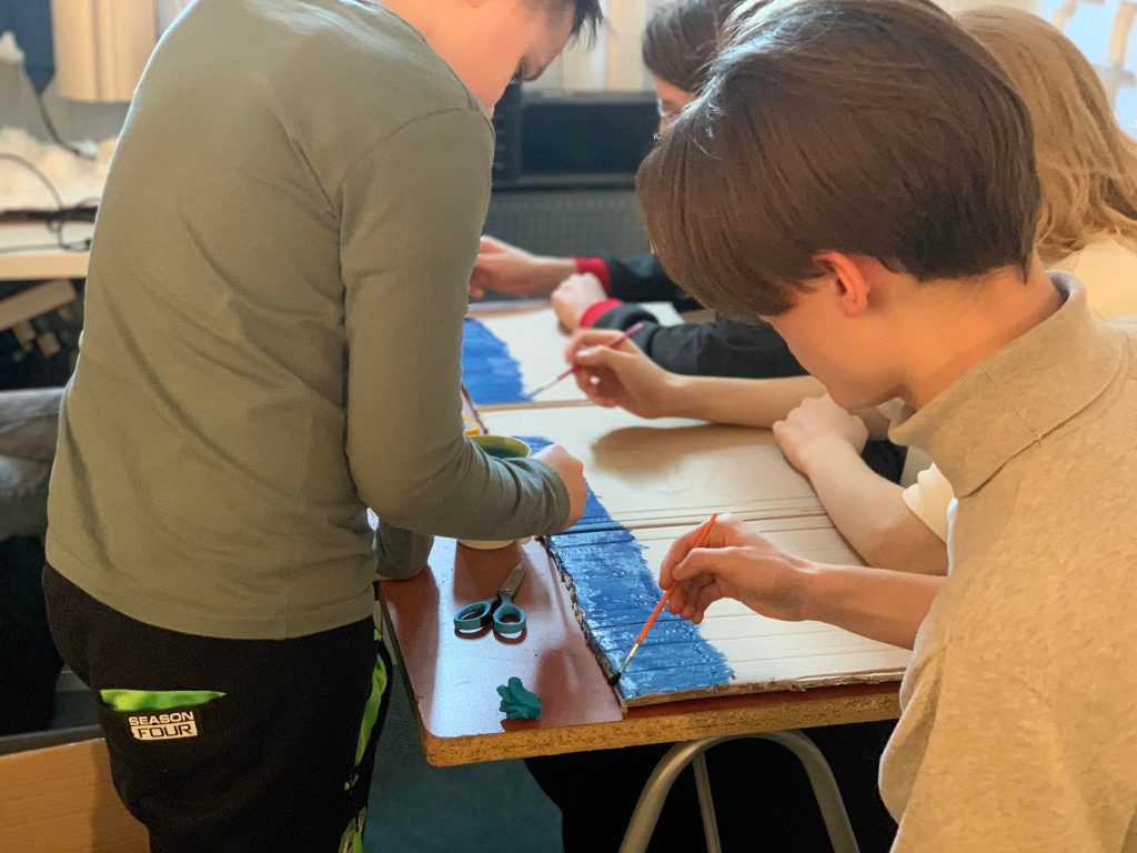 Grupa dzieci malująca na kartonie. Dzieci trzymają pędzle i malują niebieską farbą. Na stole widać nożyczki i pojemnik z farbą.