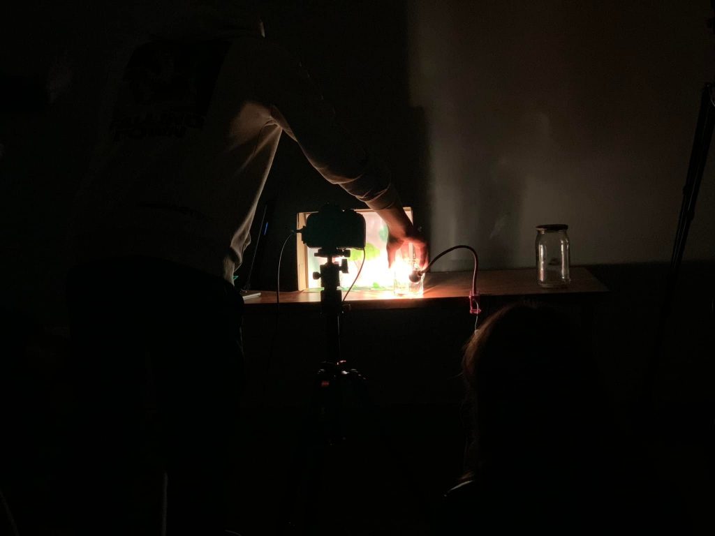 Na zdjęciu widać ciemne pomieszczenie, w którym ktoś pracuje przy stole. Osoba ta manipuluje urządzeniem, które wydaje się być kamerą lub projektorem, skierowanym na podświetloną scenkę.