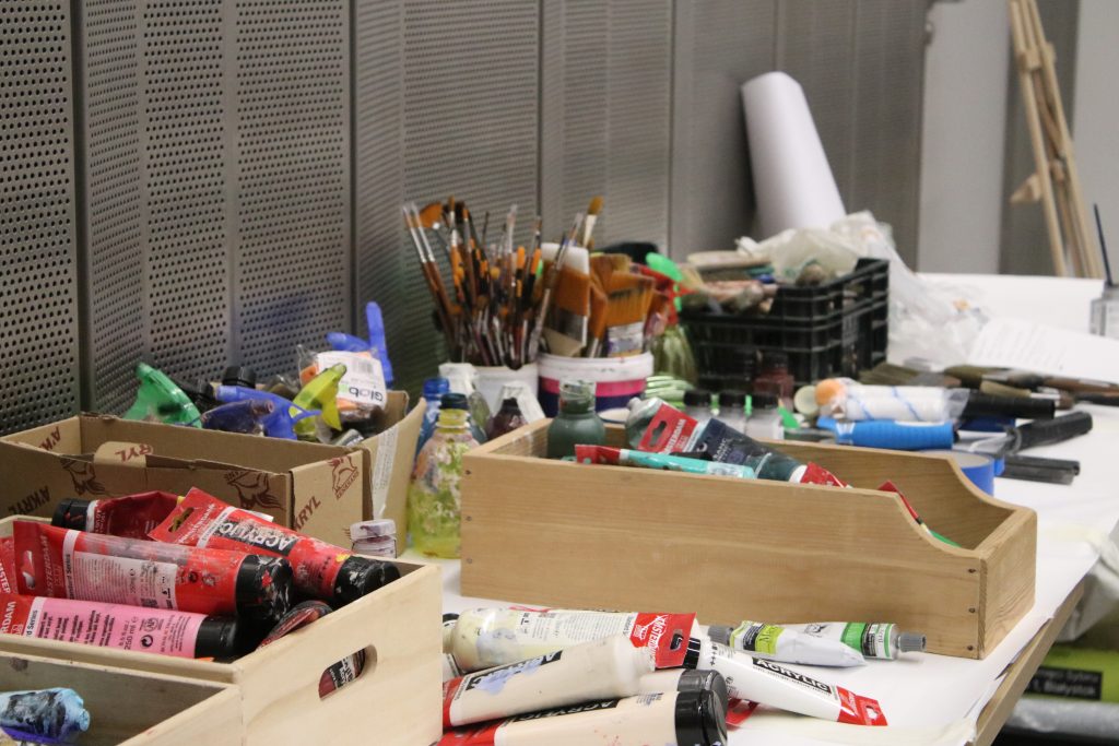 Stolik w pracowni artystycznej pełen materiałów artystycznych: drewniane i plastikowe pudełka z tubkami farb akrylowych, pędzle w kubkach, butelki z pigmentami i akcesoria malarskie.