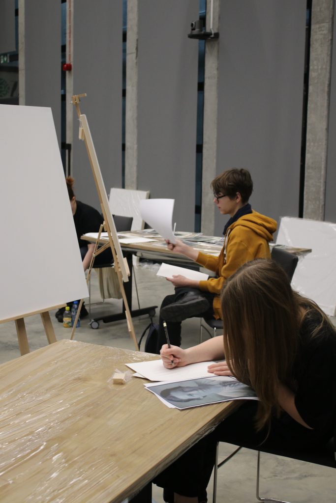 Chłopiec i dziewczynka siedzą przy stołach, rysując na kartkach. W tle widoczne są sztalugi z białymi płótnami.