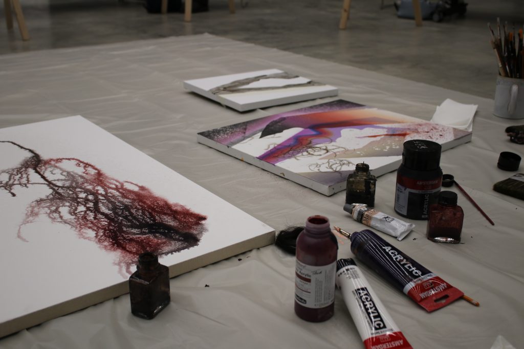 Na podłodze rozłożone są płótna z abstrakcyjnymi pracami w odcieniach czerwieni, fioletu i czerni, otaczają je butelki z farbami, tubki z akrylem, pędzle i inne narzędzia artystyczne.