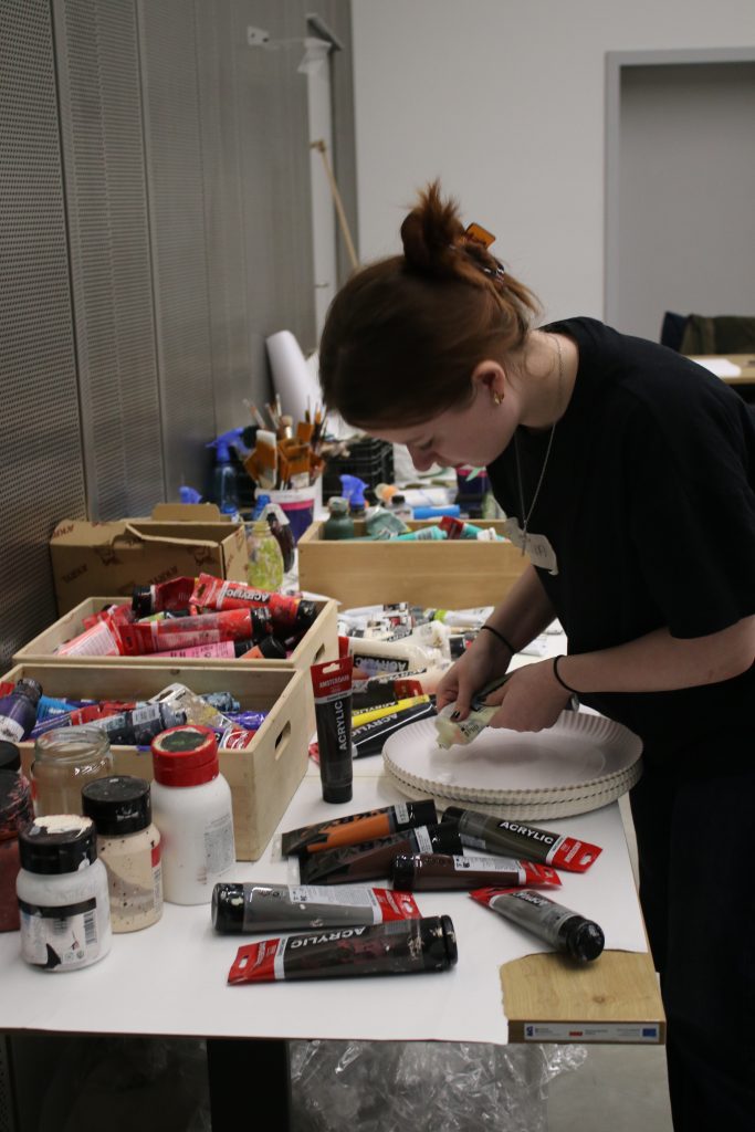 Młoda kobieta stoi przy blacie i wyciska z tubki farbę na papierowy talerz. Na blacie leżą tubki z farbami akrylowymi, pędzle, słoiki z pigmentami i pudełka z narzędziami.