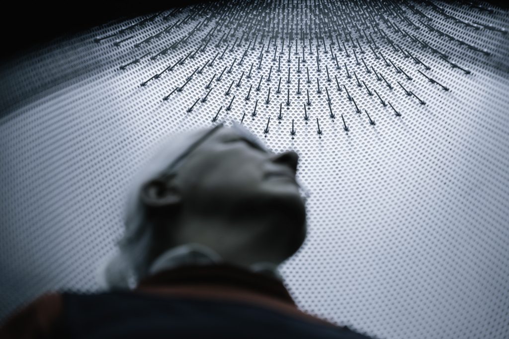 Starsza kobieta z siwymi włosami, która patrzy w górę. Znajduje się wewnątrz instalacji artystycznej, na górze której znajdują się kolce lub gwoździe.