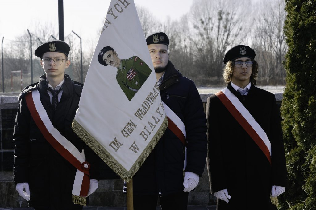 Zdjęcie przedstawia trzech młodych mężczyzn w mundurach, stojących na zewnątrz. Dwóch z nich nosi biało-czerwone szarfy, a środkowy trzyma flagę z wizerunkiem generała.
