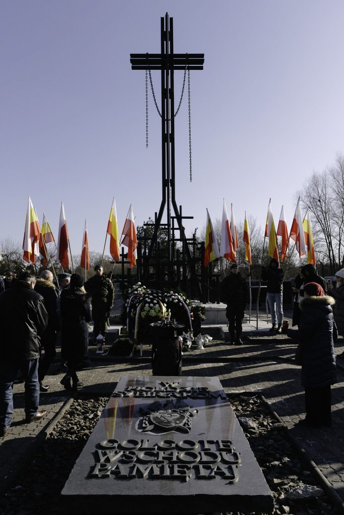 Zdjęcie przedstawia grupę ludzi przy pomniku z dużym krzyżem, otoczonym polskimi flagami. Wokół krzyża są wieńce i kwiaty.