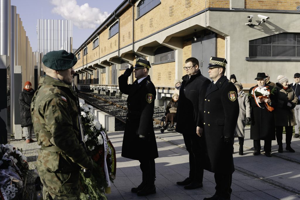 Tomasz Klim, zastępca prezydenta Białegostoku składa hołd pod pomnikiem. Obok niego stoi dwóch funkcjonariuszy straży miejskiej. W tle inni uczestnicy wydarzenia.