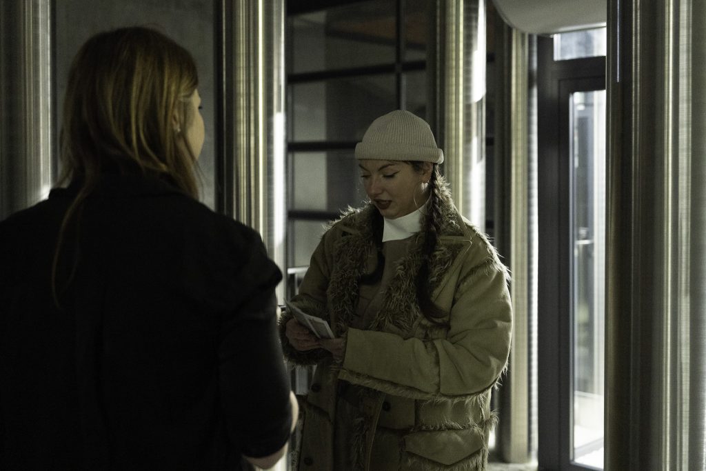 Zdjęcie przedstawia dwie osoby rozmawiające ze sobą. Jedna osoba jest odwrócona plecami do kamery, ubrana na ciemno, podczas gdy druga osoba nosi jasną czapkę, futro na kołnierzu płaszcza i trzyma jakieś papiery lub broszurę. 