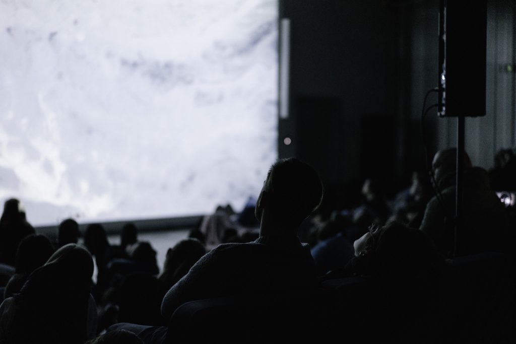 Grupa ludzi siedząca w ciemnej sali, oglądająca projekcję na dużym ekranie. 