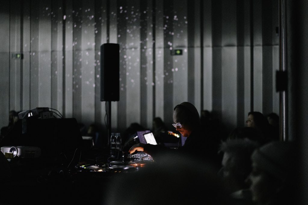 DJ w ciemnym pomieszczeniu, obok niego duży głośnik, publiczność w tle. Muzyk przy konsoli, widownia z tyłu. DJ w akcji, publiczność obserwuje. Występ DJ-a w ciemnej sali z dużym głośnikiem.