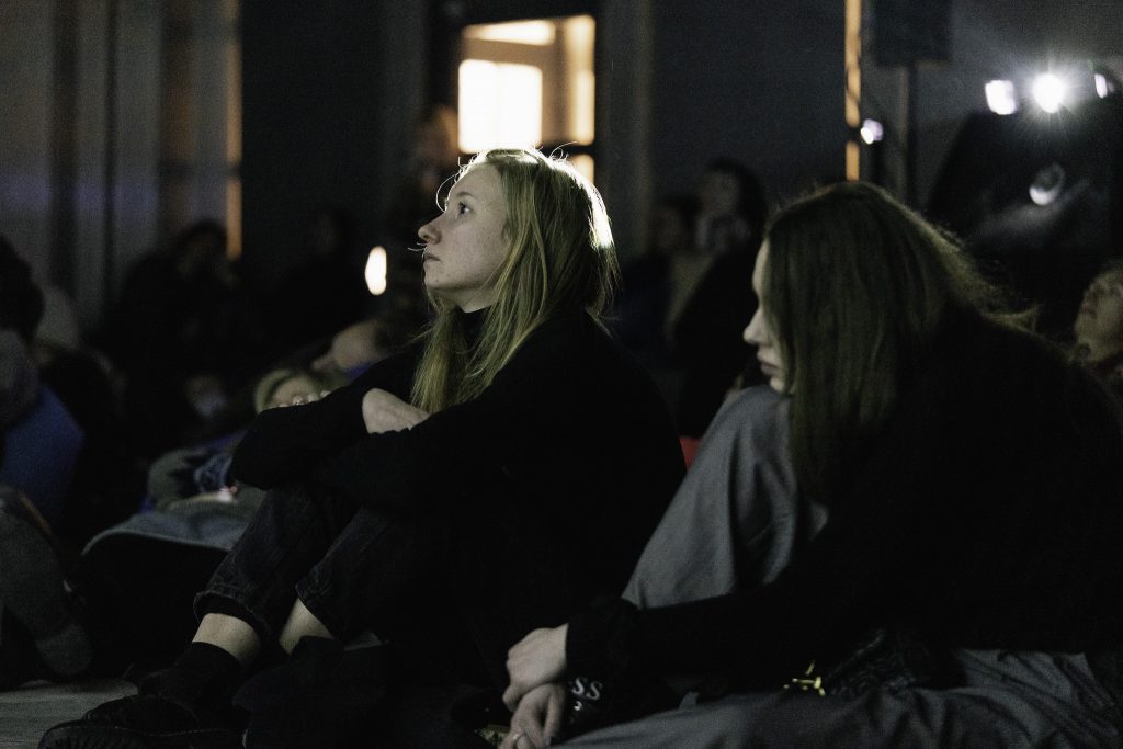 Dwie młode kobiety siedzą na podłodze w ciemnym pomieszczeniu, oglądając coś w oddali. Pomieszczenie jest słabo oświetlone, a w tle widać inne osoby. Jedna kobieta ma długie blond włosy, a druga długie ciemne włosy. Obie są ubrane w ciemne ubrania i siedzą blisko siebie, z nogami podkulonymi.