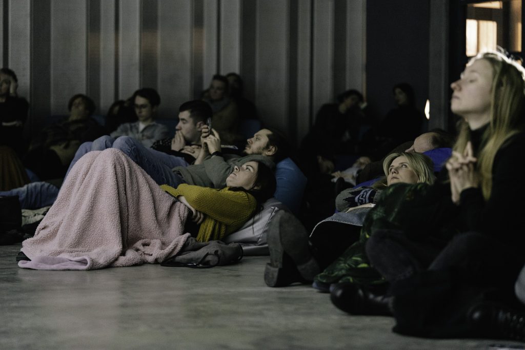 Grupa ludzi siedzi lub leży na podłodze, niektórzy są owinięci kocami.