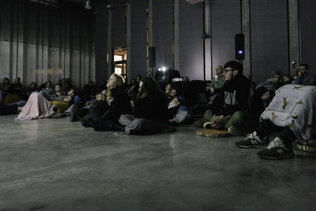Grupa ludzi siedzi lub leży na podłodze, niektórzy są owinięci kocami.