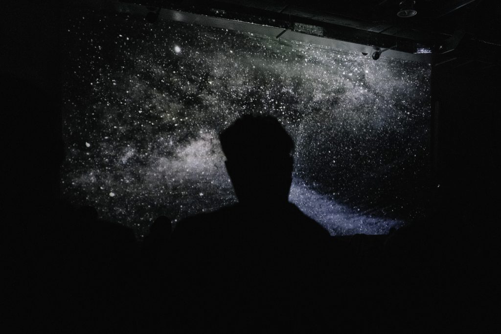 Osoba siedzi w ciemnym pomieszczeniu, obserwując projekcję na dużym ekranie. Na ekranie wyświetlane jest zdjęcie przypominające galaktykę lub kosmos z gwiazdami i mgławicami. Sylwetka osoby jest ciemna, co kontrastuje z jasnym obrazem na ekranie.
