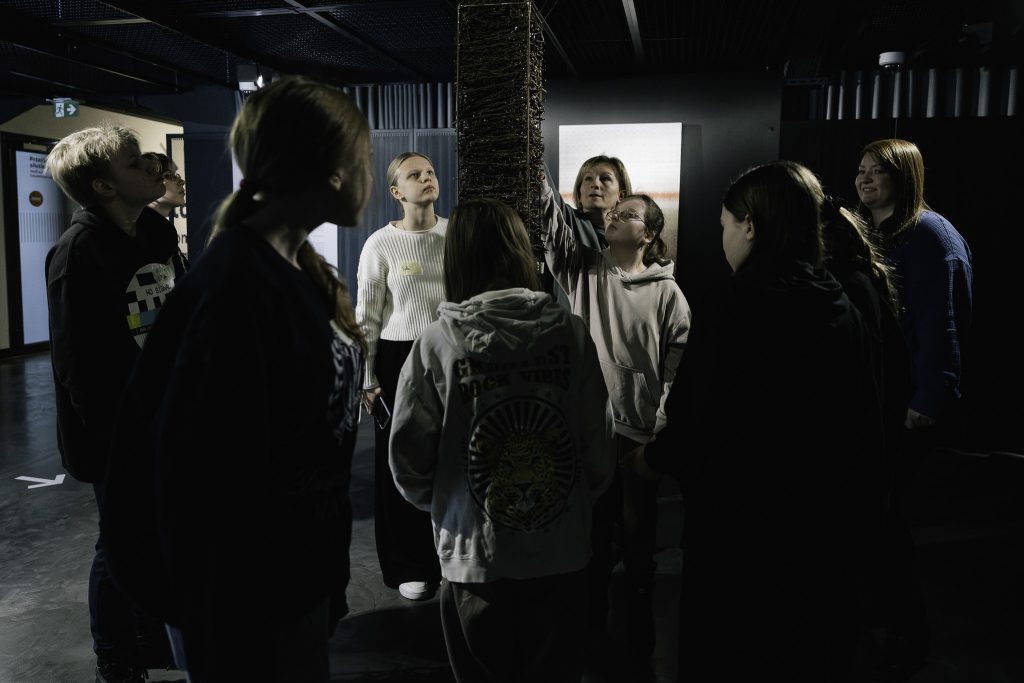 Grupa osób stoi w kręgu wokół wysokiej, pionowej instalacji artystycznej. Dwie osoby wskazują na górę tej instalacji.