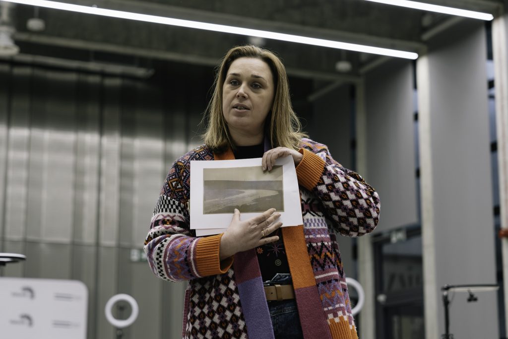 Kobieta trzyma przed sobą kartkę papieru z rysunkiem lub zdjęciem pejzażu. Ubrana jest w kolorowy, wzorzysty sweter.