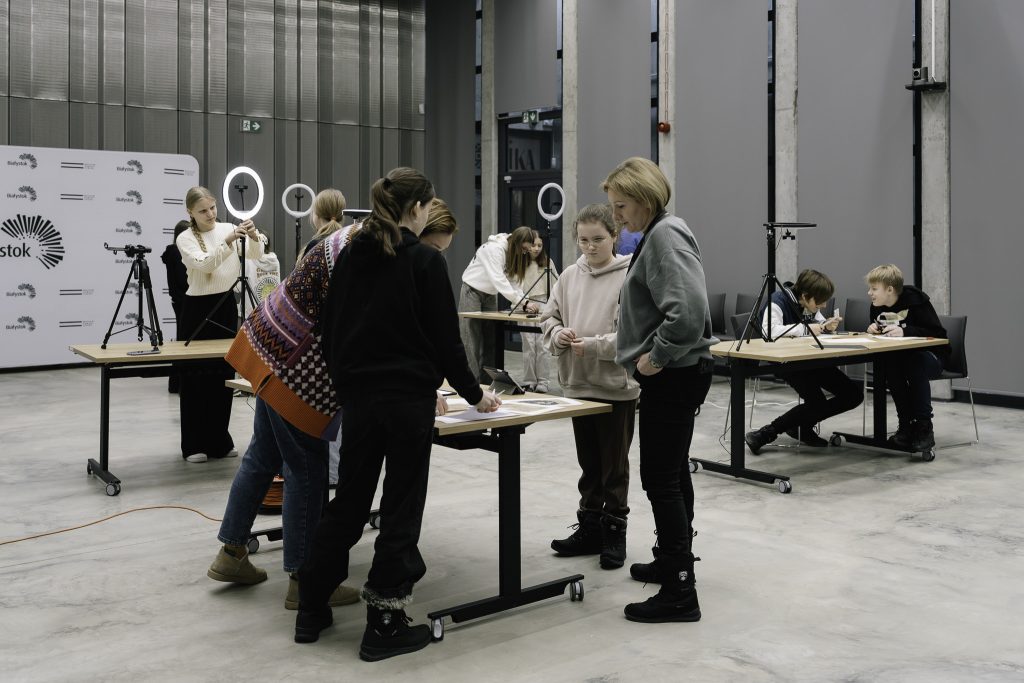 Grupa osób uczestniczy w warsztatach. Pracują przy stołach z materiałami, rozstawione są też statywy.