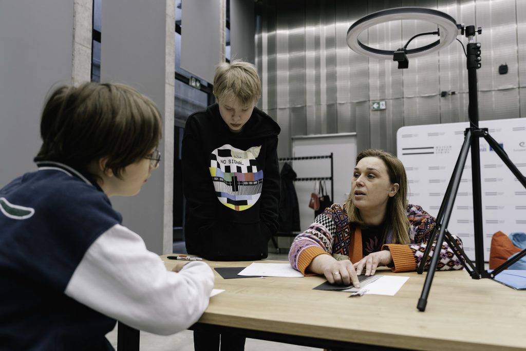 Trzy osoby siedzą przy stole, uczestnicząc w warsztatach. Na stole leżą kartki papieru i markery, a obok stoi statyw z lampą.