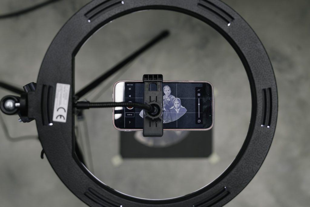 Na zdjęciu widać telefon komórkowy zamocowany na statywie z lampą. Ekran telefonu pokazuje aplikację do edycji zdjęć z nałożoną siatką, która pomaga w kadrowaniu lub edycji.