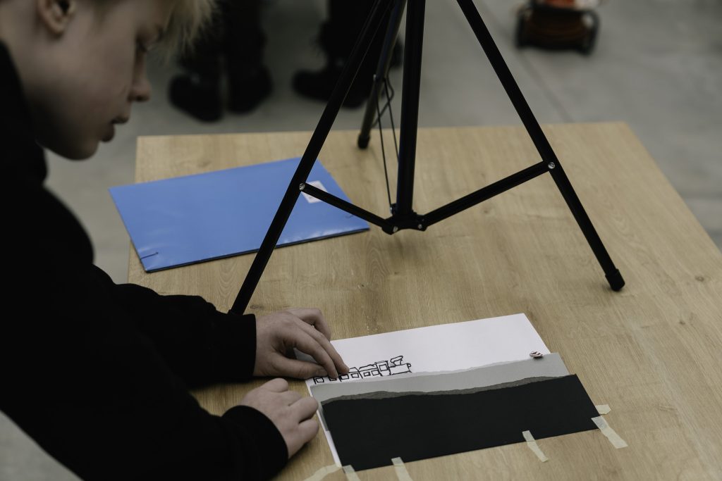 Chłpiec siedzi przy stole, pracując nad projektem artystycznym. Na stole leży kartka papieru z rysunkiem, częściowo zakryta czarnym papierem. Obok znajduje się niebieska teczka, a nad stołem stoi statyw, prawdopodobnie do oświetlenia lub nagrywania pracy.