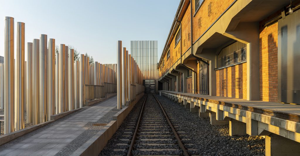 Widok na główne wejście do Muzeum. Głównym motywem są  tory biegnące w stronę budynku. Po lewej stronie widoczny symboliczny las syberyjski. Po prawej stronie widzimy odrestaurowany stary budynek magazynu, zaadaptowany obecnie na potrzeby wystawy stałej w Muzeum.