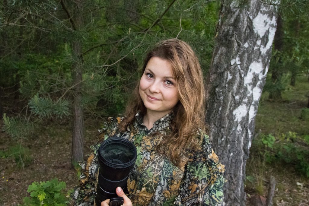 Barbara Kędzior-Adamska. Jest to młoda kobieta z długimi włosami. Trzyma w rękach duży obiektyw do aparatu fotograficznego. jest ubrana w kolorową bluzę. Stoi na tle brzozy. W głębi widać zarośla leśne.