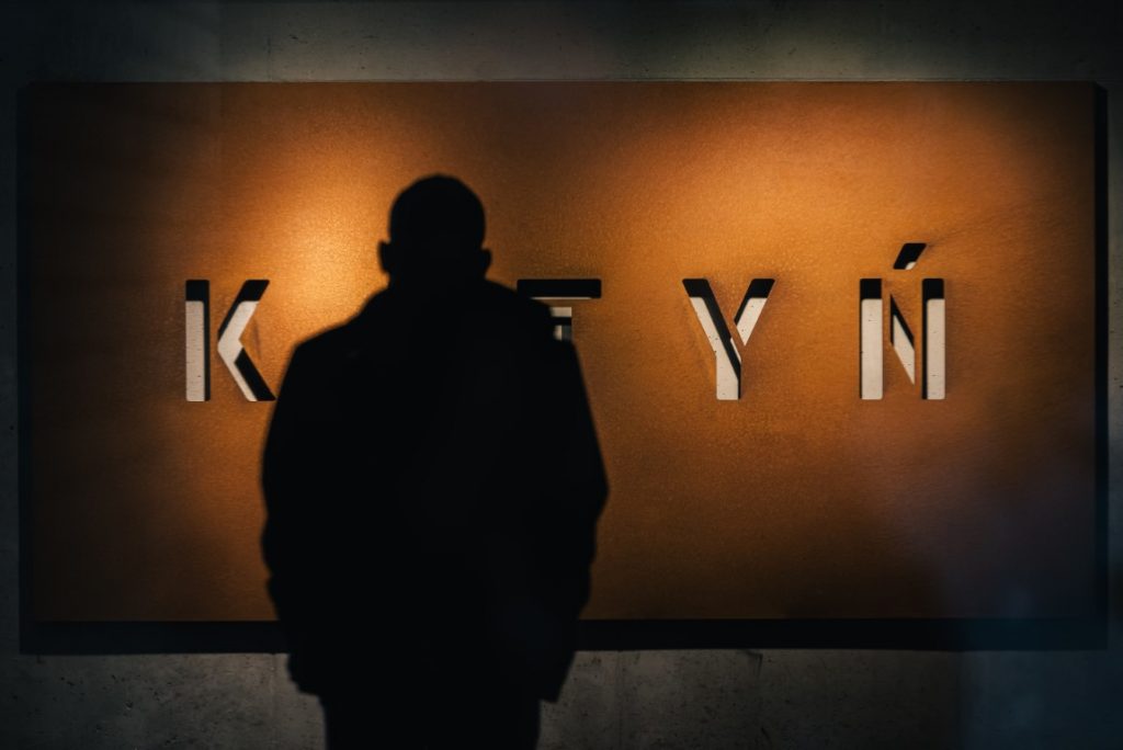 Na pierwszym planie widać ciemną sylwetkę mężczyzny patrzącego na rdzawą ścianę z napisem KATYŃ.