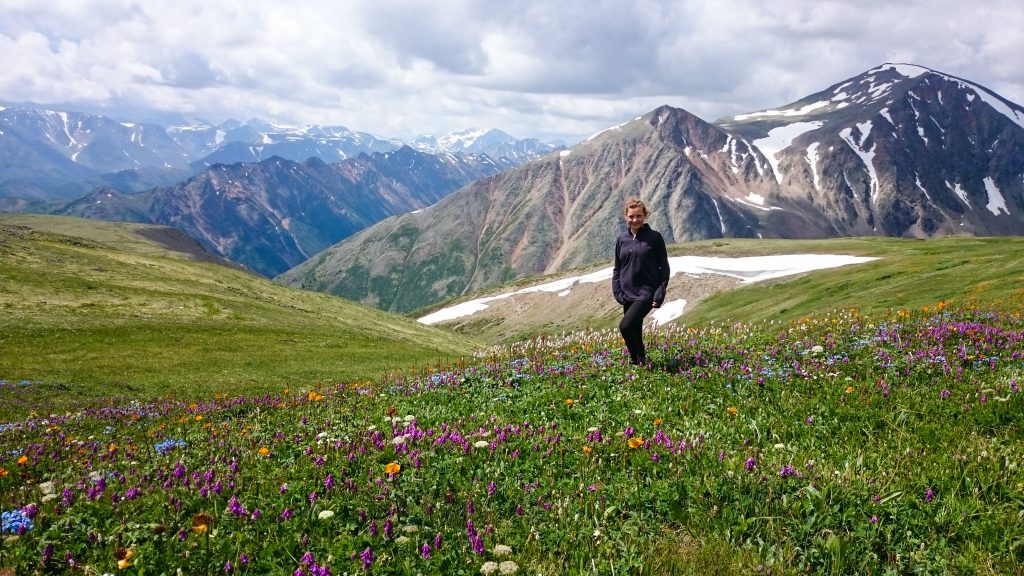 Młoda kobieta stoi na ukwieconej górskiej łące. W tle widać wysokie góry.
