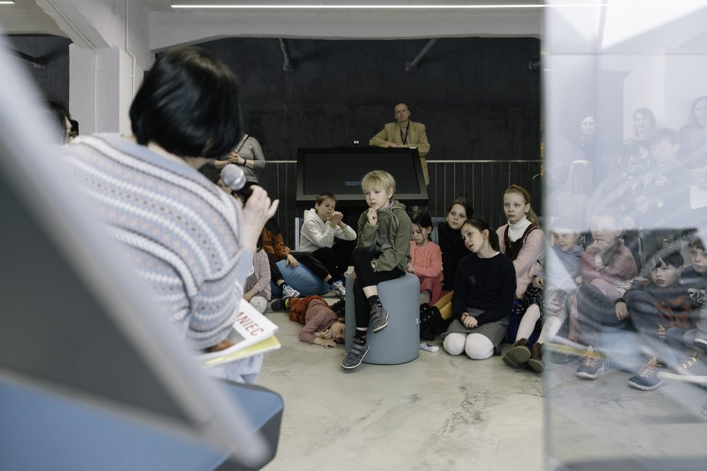 Wnuczka Heleny Pałki siedzi na fotelu. Trzyma mikrofon i przemawia do zgromadzonej publiczności. 