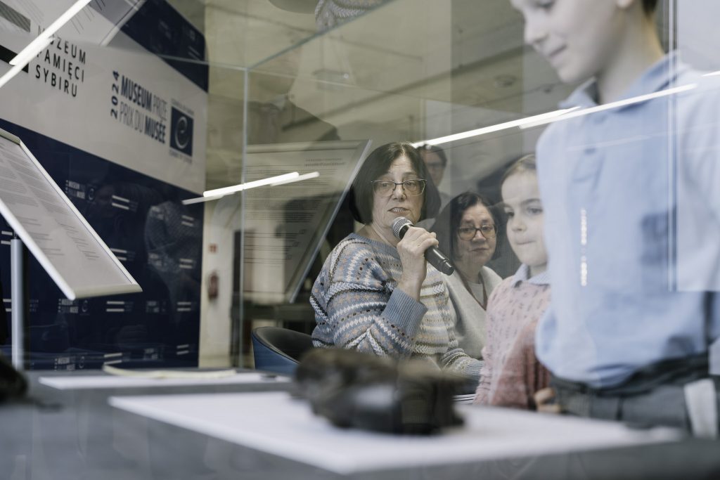 Wnuczki Heleny Pałki siedzą na fotelu. Jedna z nich trzyma mikrofon i przemawia do zgromadzonej publiczności. Spogląda na gablotę. Przy gablocie stoi dwójka dzieci.