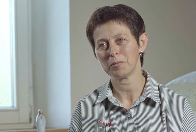 Helena Grzybkowska, starsza osoba podczas udzielania wywiadu.  Jest ubrana w koszulę w biało-czarne paski. W tle widać fragment białej ściany i okna.