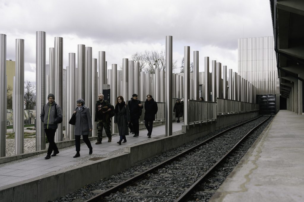 Uczestnicy Komisji Syberyjskiej wychodzą z gmachu Muzeum. Idą w stronę pomnika Matki Sybiraczki. Widać tory biegnące w stronę budynku. W tle widoczny jest las metalicznych słupów.