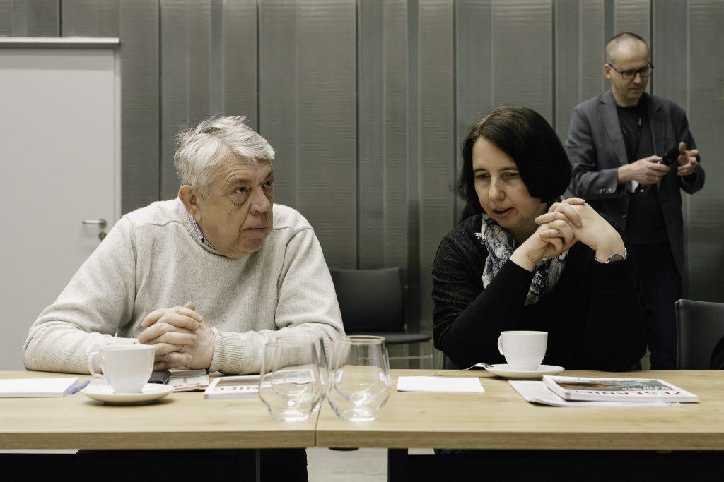 Dwoje uczestników Komisji Syberyjskiej siedzą przy stole. Na stole stoją szklanki, filiżanki, książki. Za nimi, w tle widoczny jest dr Marcin Zwolski.