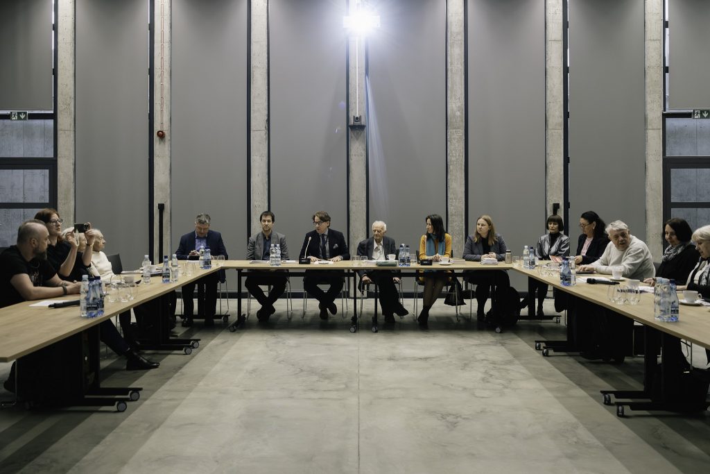 Uczestnicy Komisji Syberyjskiej siedzą przy stole. Na stole znajdują się różne przedmioty, butelki z wodą, szklanki, materiały konferencyjne, mikrofony.