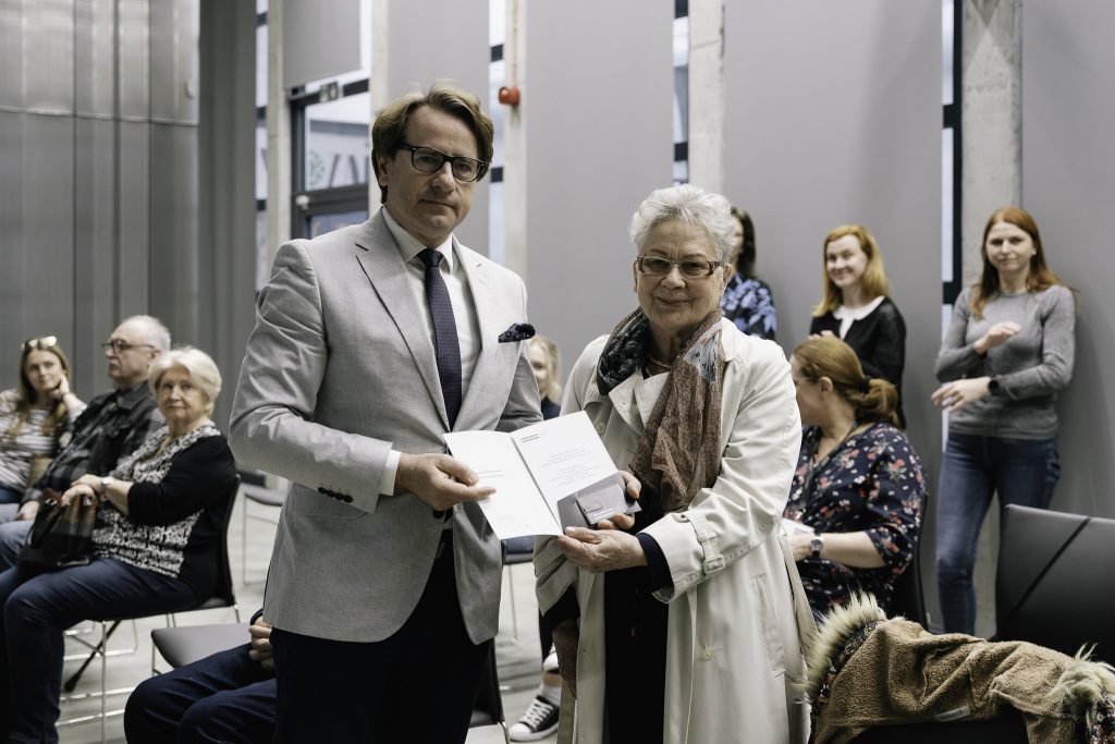 Dyrektor Muzeum Pamięci Sybiru, prof. Wojciech Śleszyński wręcza dla starszej kobiety, Sybiraczki, podziękowania za podzielenie się własną historią. W tle grupa osób siedzi na krzesłach.