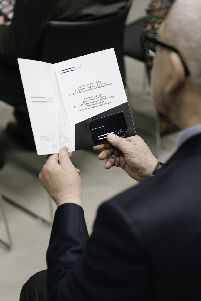 Starszy mężczyzna w garniturze trzyma w rękach podziękowania za podzielenie się własną historią.