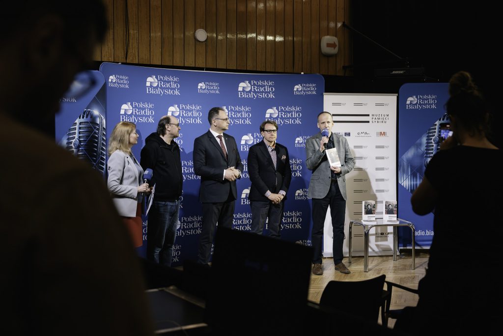 Grupa kilku osób podczas konferencji prasowej. Od lewej stoją Adam Woronowicz, aktor filmowy i teatralny,  Marcin Tomkiel, redaktor naczelny Polskiego Radia Białystok, prof. Wojciech Śleszyński, dyrektor Muzeum Pamięci Sybiru, dr Marcin Zwolski, kierownik działu naukowego Muzeum Pamięci Sybiru. Dr Marcin Zwolski trzyma w ręku mikrofon i przemawia. W drugiej ręce trzyma książkę Romualda Lipińskiego „Życiorys pisany przez historię”. 