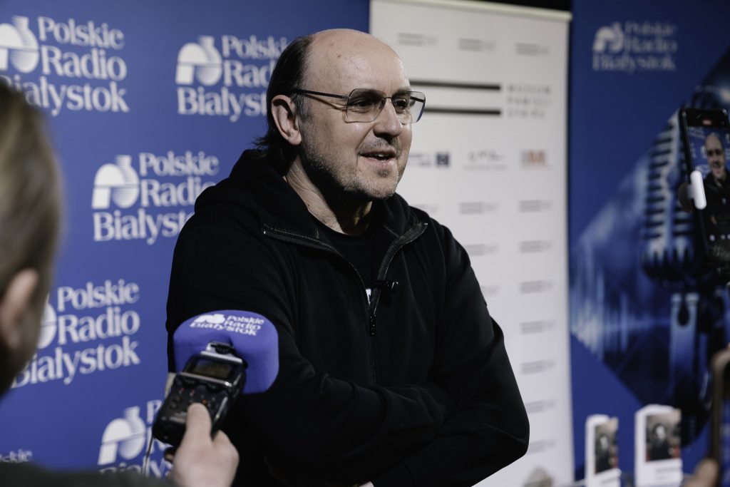 Adam Woronowicz, aktor filmowy i teatralny udziela wywiadu. Jest to mężczyzna w średnim wieku w okularach. Dziennikarka trzyma w ręku dyktafon z logo Polskiego Radia Białystok.