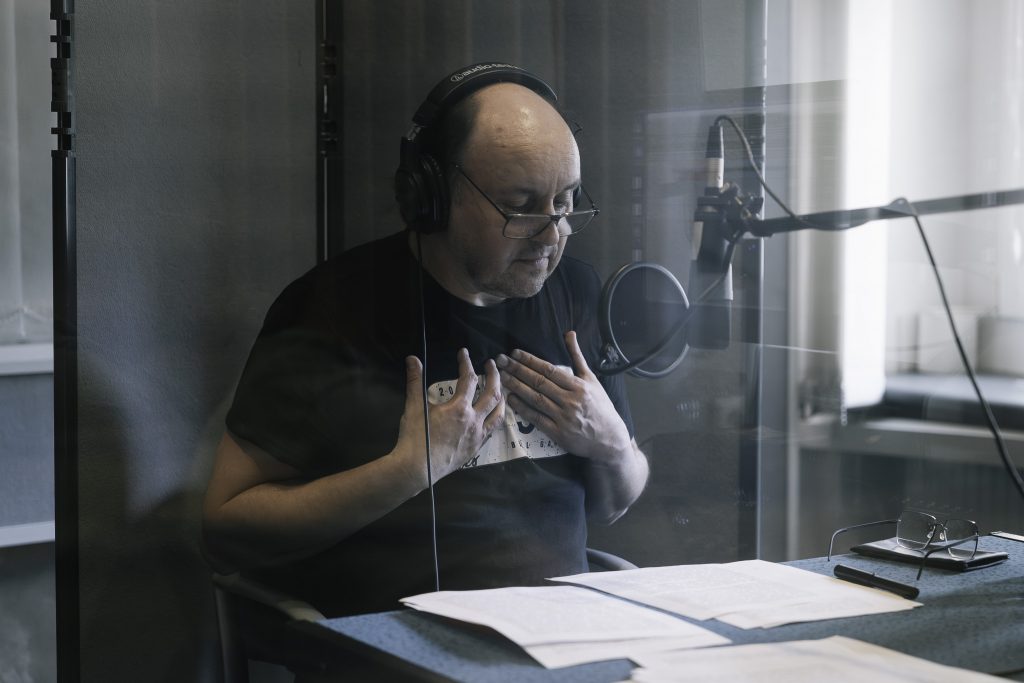 Adam Woronowicz podczas nagrania audiobooka. Siedzi w profesjonalnym studio nagraniowym. Przed nim jest mikrofon.