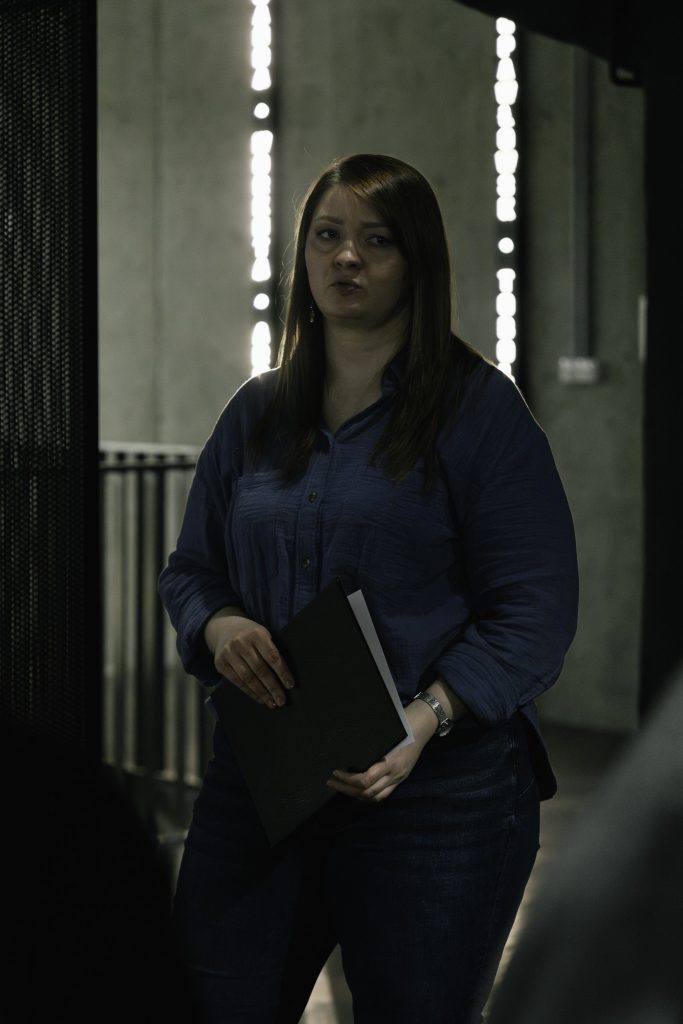 Kuratorka wystawy, Ewelina Kamieńska. jest ubrana w ciemnoniebieskie spodnie i bluzkę. W rękach trzyma ciemną teczkę. W tle widoczna betonowa ściana z podświetlonymi nazwami miejscowości do których byli zsyłani Sybiracy.