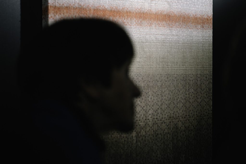 Sylwetka jednej z osób zwiedzających wystawę. W tle widoczny jeden z eksponatów, ręcznie tkane płótno.