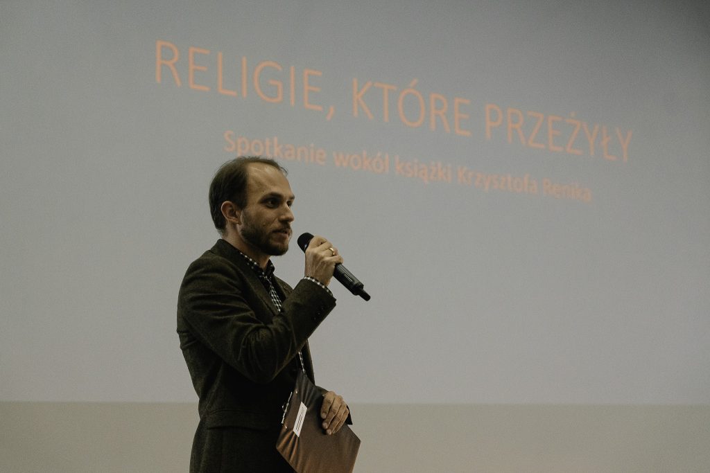 Dr Piotr Popławski, kierownik Działu Projektów Kulturalnych Muzeum Pamięci Sybiru przemawia podczas spotkania. Jest ubrany w ciemnobrązową marynarkę, trzyma w ręku mikrofon. W tle widoczny kadr z wyświetlanej prezentacji, czerwony napis "Religie, które przeżyły. Spotkanie wokół książki Krzysztofa Renika" na czarnym tle.